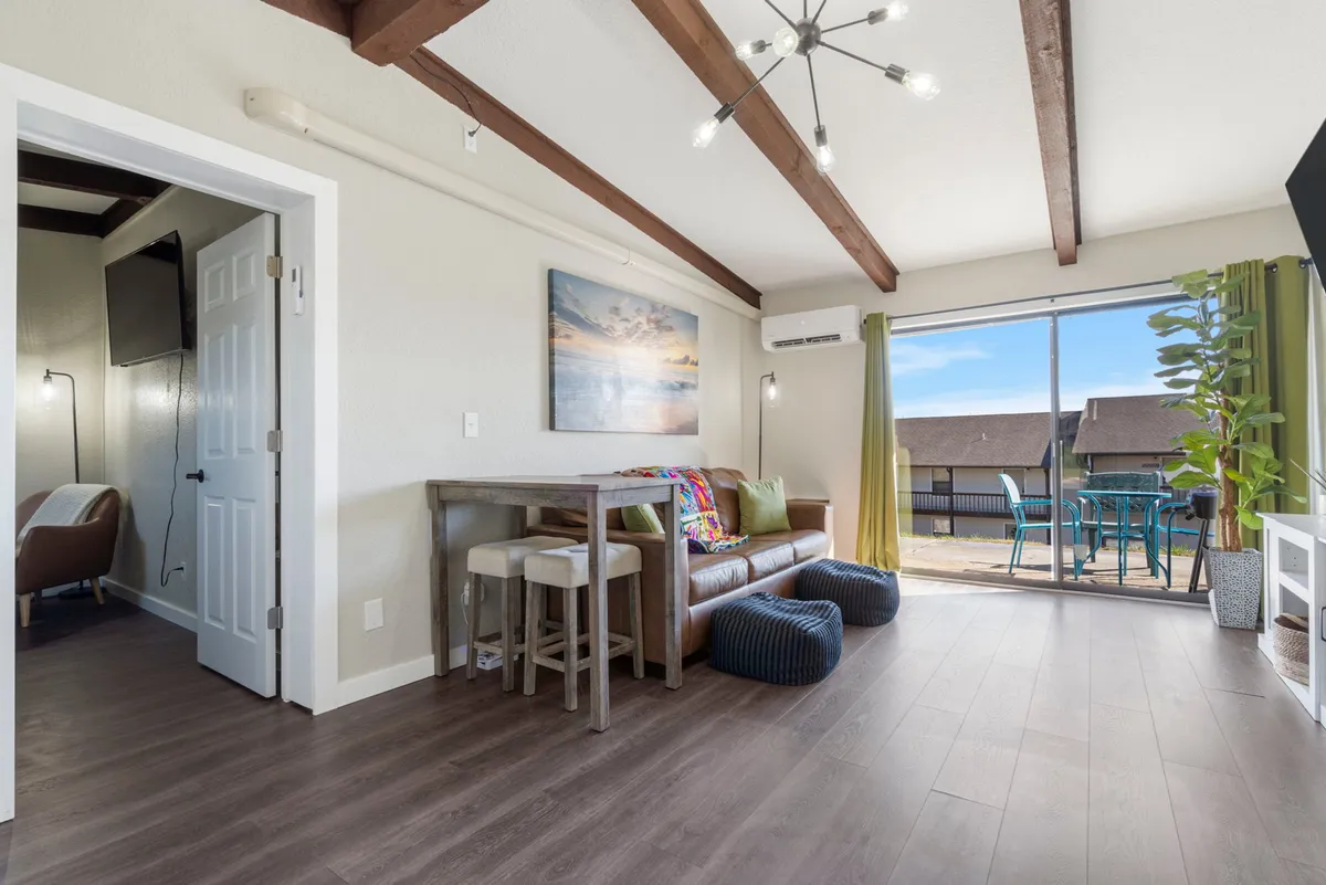 A cozy living area featuring a plush sofa with a colorful throw, large glass doors that open to an outdoor space, and inviting artwork on the wall. Morning light fills the room, creating a welcoming and relaxing atmosphere.