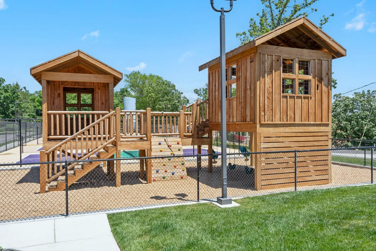 This image captures a lively children's playground with wooden playhouses, a climbing structure, and various playground equipment set amongst greenery. The scene is filled with opportunities for imaginative play and socializing, radiating a joyful, family-friendly mood—perfect for families with young children.