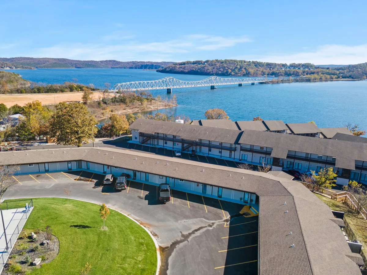 A sweeping exterior shot showcasing the tranquil lakeside with Table Rock Lake in the foreground, the iconic Kimberling City Bridge arching across the water, and a welcoming hotel structure nestled in lush green lawns. Boaters and early morning light hint at a peaceful, romantic atmosphere ideal for couples.