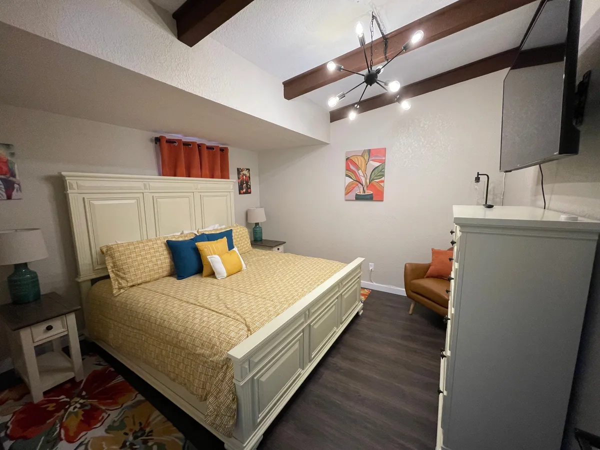 A welcoming bedroom featuring a neatly made bed with decorative pillows, tasteful artwork on the wall, a warm chandelier overhead, and comfortable furniture. The room exudes a sense of calm and privacy, ideal for relaxation and unwinding.