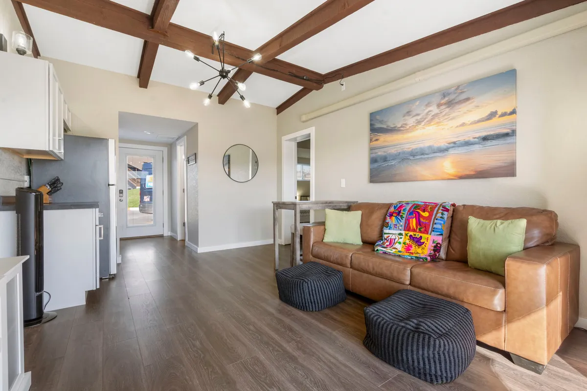 A warm, cozy living area with a kitchen in the background, leather sofa adorned with colorful throw pillows, and a dining table set for a meal. Beach sunset artwork adds a vacation vibe, and the layout encourages socializing and shared meals.
