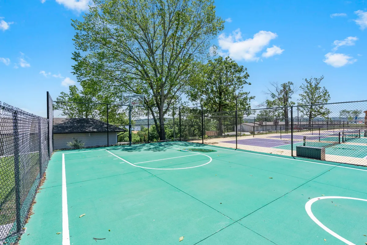 An energetic outdoor basketball court surrounded by trees, featuring a prominent basketball hoop and a vivid green court surface. The open area invites groups for friendly games or tournaments, with a backdrop of natural greenery for atmosphere.
