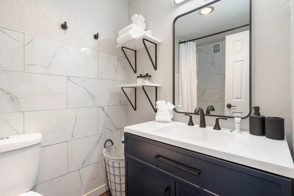 A contemporary bathroom with elegant marble wall tiles, stylish black faucet fixtures, a large mirror reflecting ample light, and neatly stacked towels. The mood is calming and sophisticated, inviting guests to enjoy moments of self-care.