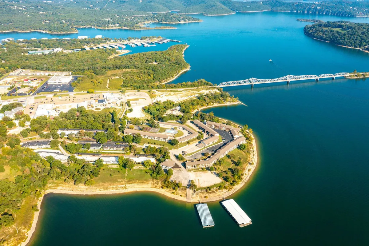 A broad view across Table Rock Lake, featuring the lake itself, multi-family housing on the shoreline, a marina with boats docked, and a nearby bridge. The scene blends elements of water-based activities and the surrounding Ozark landscape, inviting ideas of boating, fishing, and nature exploration. The lighting suggests either early morning or late evening, amplifying the sense of adventure and tranquility.
