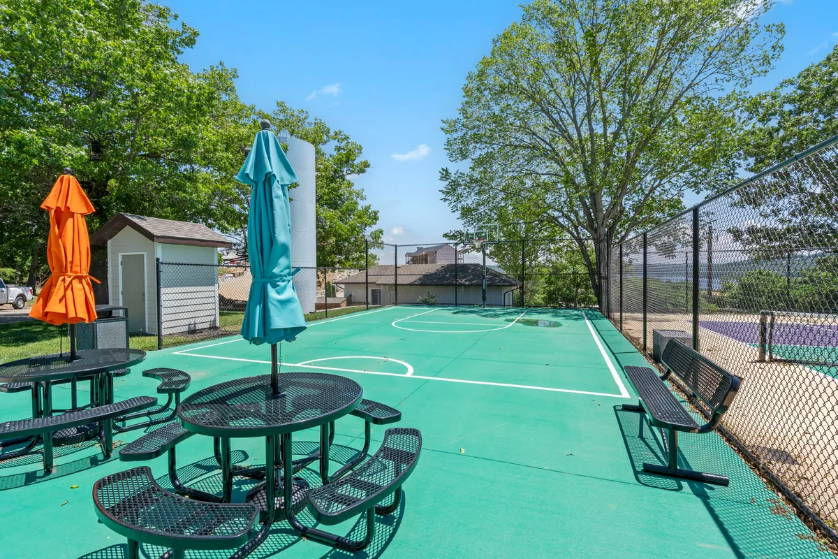 A vibrant outdoor recreation area featuring a basketball court, colorful umbrellas shading picnic tables, and lush greenery surrounding the space. The setting is lively and inviting, perfect for sports, casual socializing, or relaxing in the shade between activities.