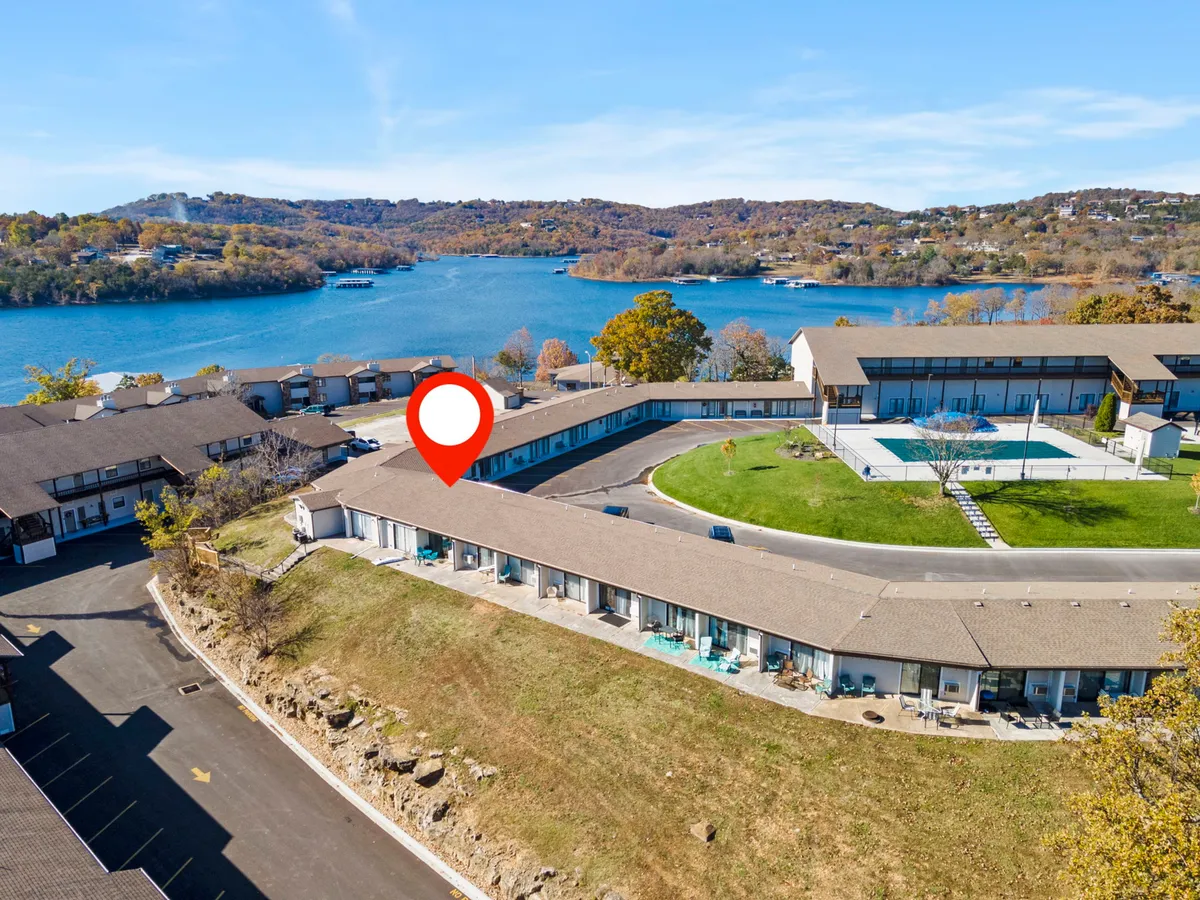 A striking exterior shot with a shimmering swimming pool in the foreground, landscaped green space, and the lake visible just beyond. The architectural style suggests a relaxed, resort-like property setting, with ample space for guests to unwind near the water.