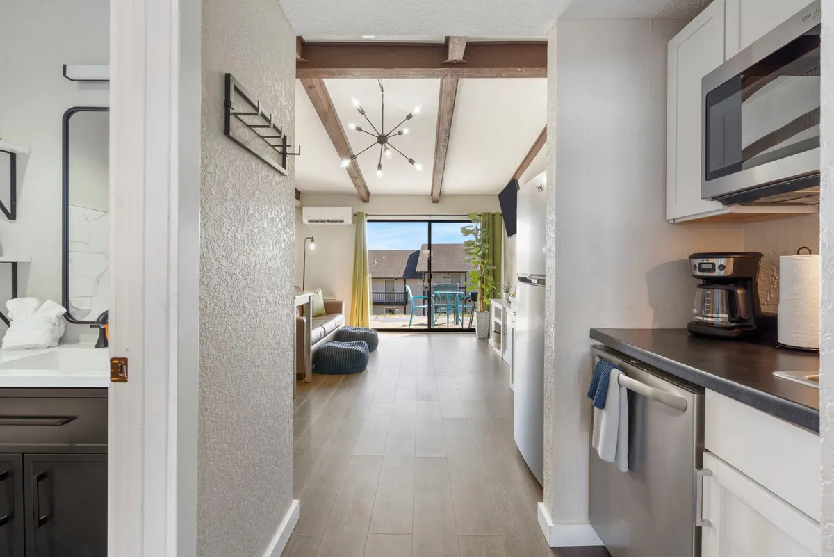 A bright, contemporary space with an open doorway leading to a stylish living area, modern kitchen appliances visible, and a clear view through glass doors to an inviting outdoor patio. The space is airy and designed for both entertaining and relaxation.
